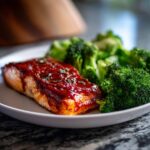 A close-up of a glistening piece of Sheet Pan Honey Harissa Salmon served next to bright green steamed broccoli florets.
