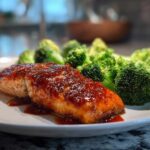 Close-up of a perfectly cooked fillet of Sheet Pan Honey Harissa Salmon topped with a glossy red glaze, served next to bright green steamed broccoli florets.