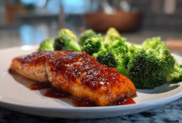 Close-up of a perfectly cooked fillet of Sheet Pan Honey Harissa Salmon topped with a glossy red glaze, served next to bright green steamed broccoli florets.