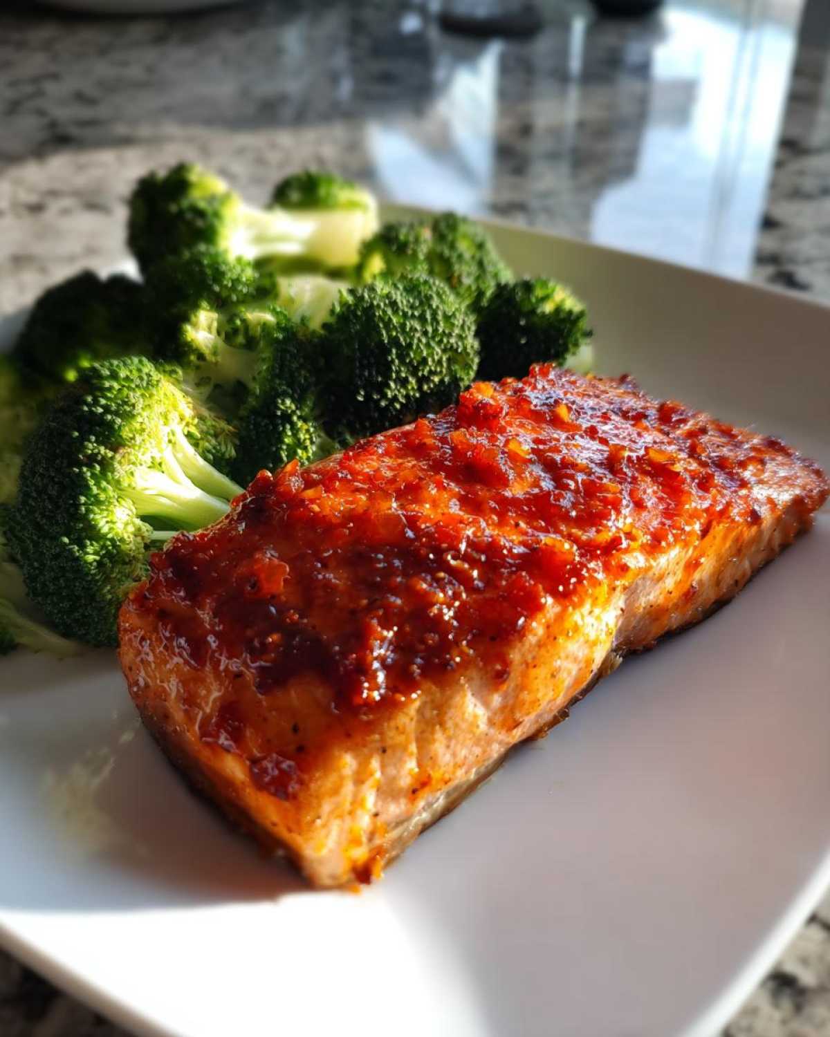 A perfectly cooked fillet of Sheet Pan Honey Harissa Salmon topped with a bright red glaze, served next to steamed broccoli florets.