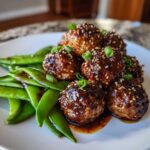 A close-up of Sheet Pan Sticky Sesame Meatballs coated in glaze and sesame seeds, served next to bright green snap peas.