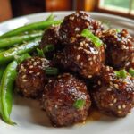Close-up of glossy Sheet Pan Sticky Sesame Meatballs coated in sauce and sesame seeds, served with bright green snap peas.