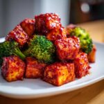 Close-up of crispy tofu cubes coated in sweet chili sauce served with bright green broccoli florets.