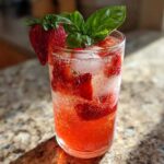 A tall glass filled with Sparkling Strawberry Basil Lemonade, ice, and garnished with a fresh strawberry and basil leaves.