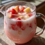 Close-up of a glass pitcher filled with pink Strawberry Piña Colada Punch, topped with chunks of strawberry and pineapple.