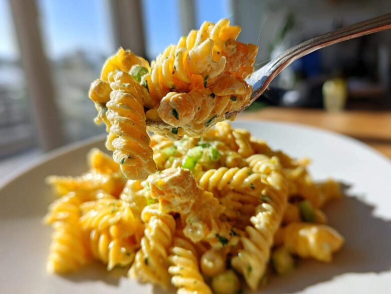 A fork lifts a bite of Vegan Creamy Lemon Chickpea Pasta Salad, showing fusilli pasta coated in a rich, yellow sauce with chickpeas.