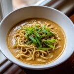 Close-up of a white bowl filled with Vegan Creamy Miso Sesame Noodle Soup, topped with green onions and sesame seeds.