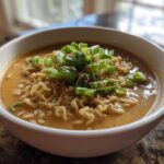A close-up of a white bowl filled with Vegan Creamy Miso Sesame Noodle Soup, topped with green onions and sesame seeds.