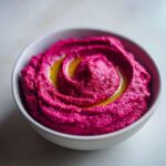 Close-up of vibrant magenta Vegan Creamy Roasted Beet Hummus swirled in a white bowl, topped with olive oil.