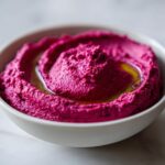 Close-up of vibrant pink Vegan Creamy Roasted Beet Hummus swirled in a white bowl, drizzled with olive oil.
