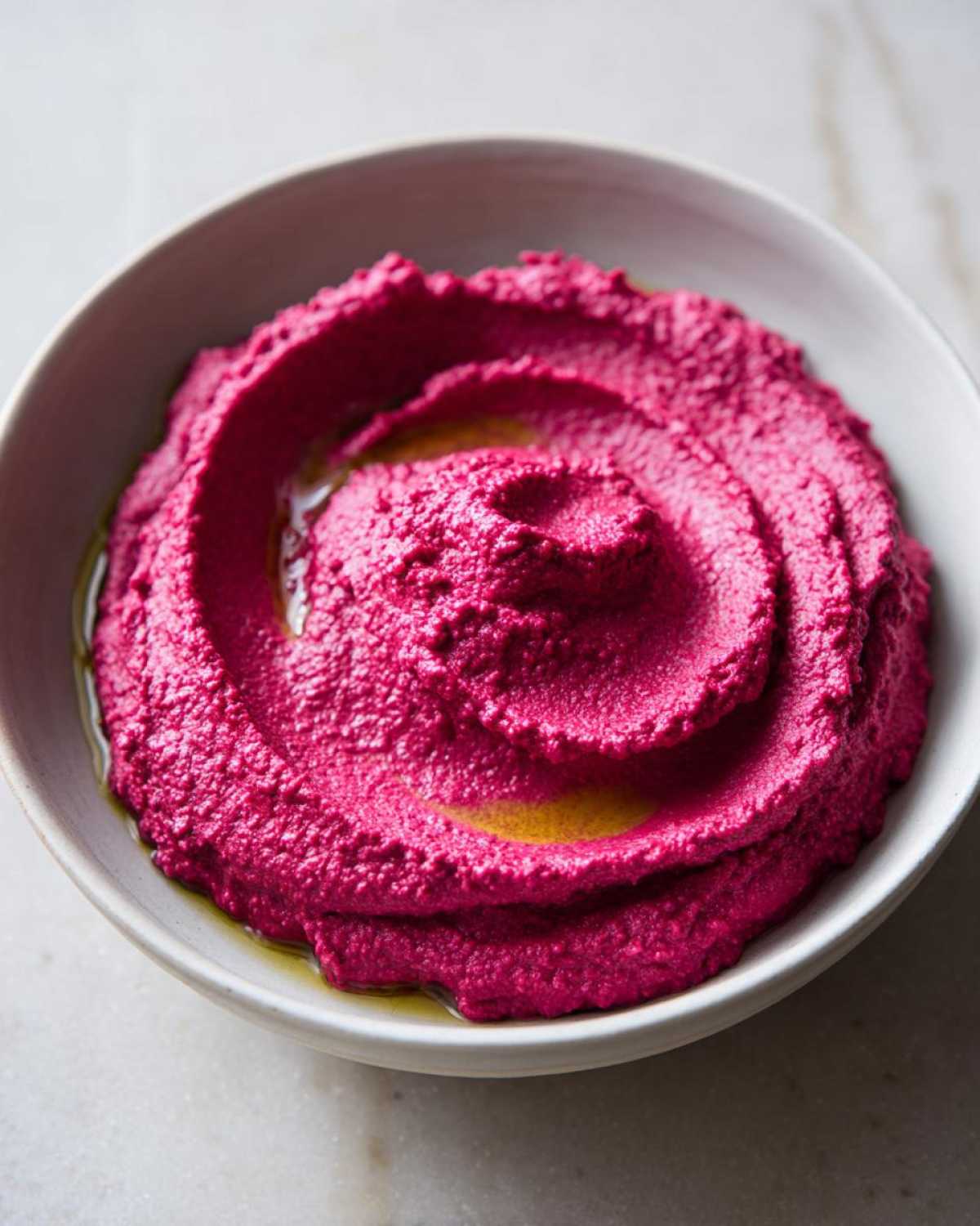 Close-up of vibrant pink Vegan Creamy Roasted Beet Hummus swirled in a white bowl and drizzled with olive oil.