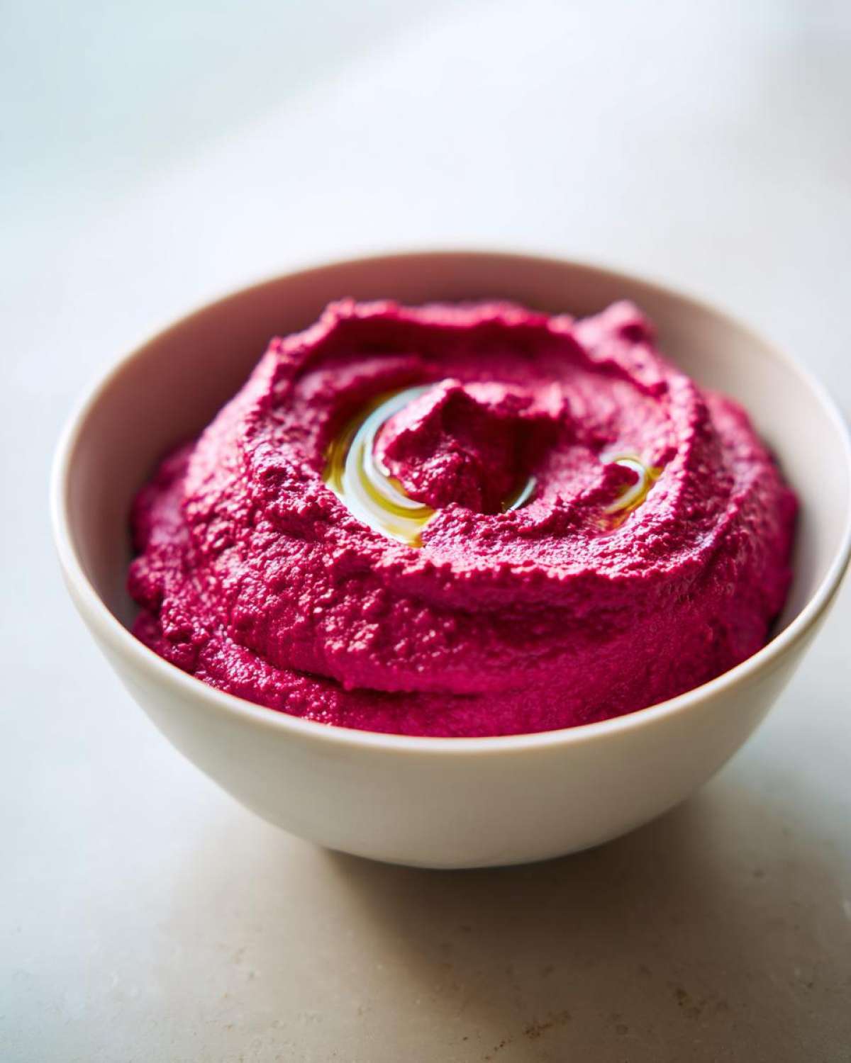 Close-up of bright magenta Vegan Creamy Roasted Beet Hummus topped with a swirl of olive oil in a white bowl.