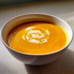 Close-up of a vibrant orange Vegan Creamy Roasted Carrot Ginger Soup topped with a swirl of white cream.