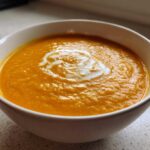 A close-up of vibrant orange Vegan Creamy Roasted Carrot Ginger Soup topped with a swirl of white cream in a white bowl.