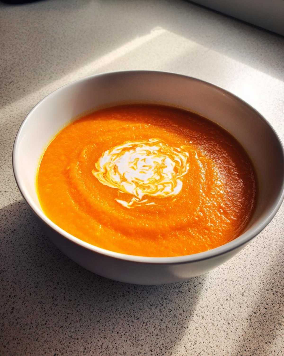 A vibrant orange bowl of Vegan Creamy Roasted Carrot Ginger Soup, swirled with a white creamy topping.