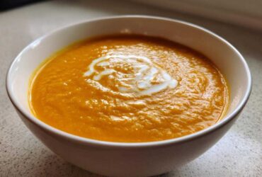 A close-up of vibrant orange Vegan Creamy Roasted Carrot Ginger Soup topped with a swirl of white cream in a white bowl.