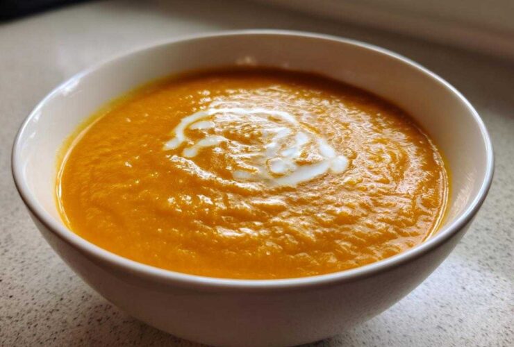 A close-up of vibrant orange Vegan Creamy Roasted Carrot Ginger Soup topped with a swirl of white cream in a white bowl.