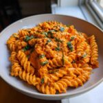 Close-up of fusilli pasta coated in a rich, orange Vegan Creamy Roasted Red Pepper Hummus Pasta sauce, topped with fresh basil.