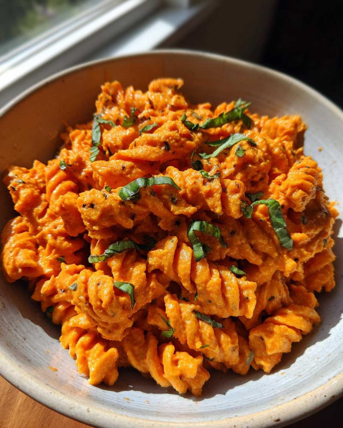 Close-up of fusilli pasta coated in bright orange Vegan Creamy Roasted Red Pepper Hummus Pasta sauce, garnished with basil.