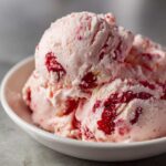 Close-up of three scoops of pink Vegan Creamy Strawberry Oat Milk Ice Cream with visible strawberry pieces in a white bowl.