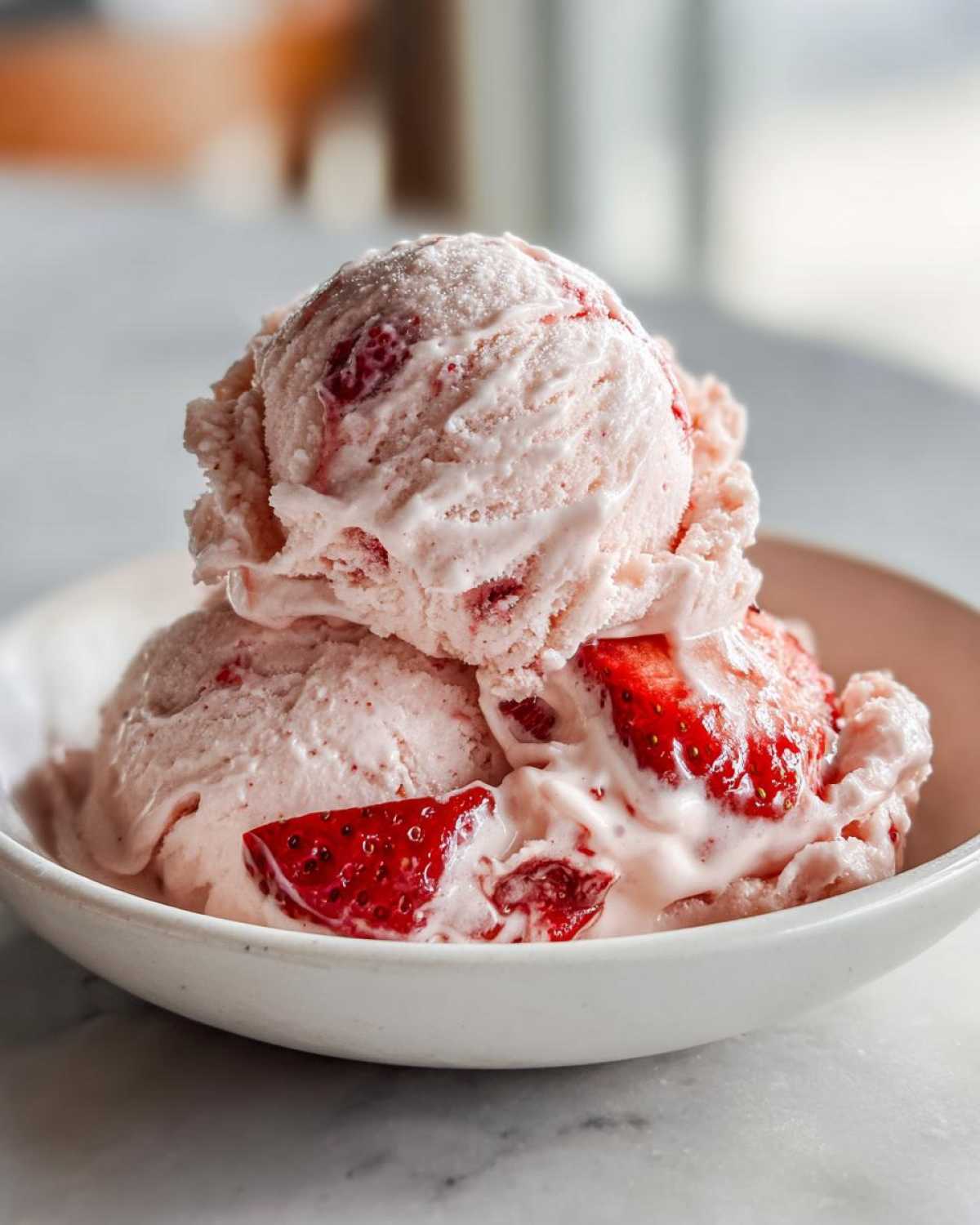 Three scoops of pink Vegan Creamy Strawberry Oat Milk Ice Cream with visible strawberry pieces in a white bowl.