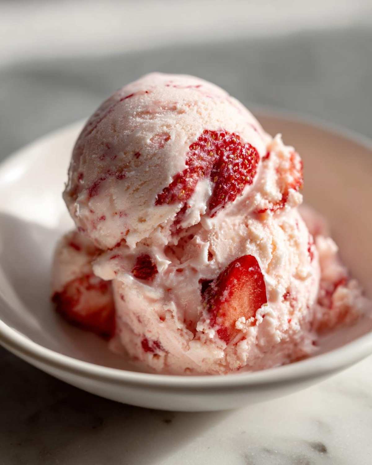Two scoops of creamy Vegan Creamy Strawberry Oat Milk Ice Cream with visible strawberry pieces in a white bowl.
