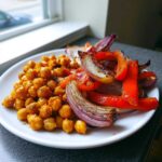 A close-up of a white plate featuring seasoned chickpeas next to roasted red peppers and red onion wedges from the Vegan One-Pan Chickpea Shawarma Sheet Bake.