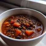 Close-up of a steaming white bowl filled with rich Vegan One-Pot Lentil Taco Soup featuring lentils, black beans, and diced orange vegetables.
