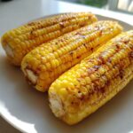 Three ears of golden yellow Air Fryer Corn on the Cob, lightly charred and seasoned, resting on a white plate.