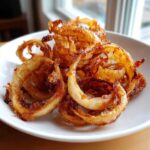 Close-up of perfectly crispy, golden-brown Air Fryer Onion Boil rings piled on a white plate.