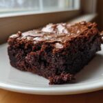 Close-up of a rich, fudgy slice of brownies with a crackly top, sitting on a white plate.