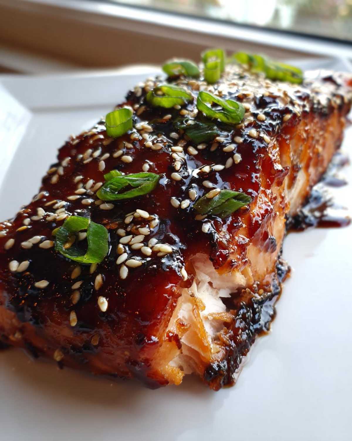 Close-up of a flaky Baked Teriyaki Salmon fillet covered in dark, glossy glaze, sesame seeds, and sliced green onions.