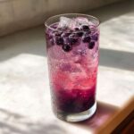 A tall, sweating glass filled with vibrant purple Blueberry Lemonade, ice cubes, and fresh blueberries.