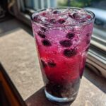 Close-up of a tall glass filled with vibrant, fizzy Blueberry Lemonade, whole blueberries, and ice cubes, sitting on a sunlit windowsill.