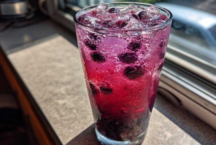 Close-up of a tall glass filled with vibrant, fizzy Blueberry Lemonade, whole blueberries, and ice cubes, sitting on a sunlit windowsill.
