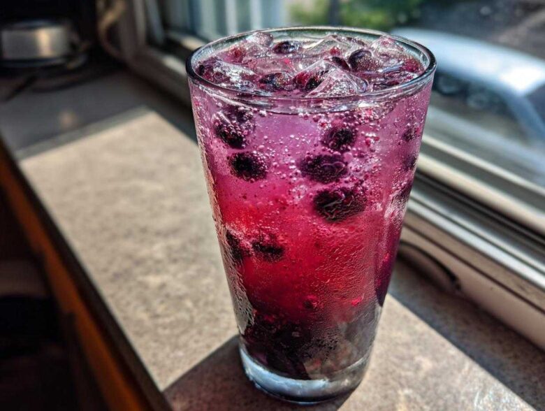 Close-up of a tall glass filled with vibrant, fizzy Blueberry Lemonade, whole blueberries, and ice cubes, sitting on a sunlit windowsill.