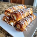 A stack of golden-brown Cinnamon Roll French Toast Roll-Ups drizzled with white icing and powdered sugar.