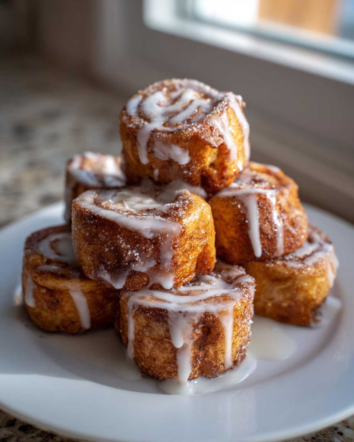 A stack of golden brown Cinnamon Roll French Toast Roll-Ups drizzled generously with white icing on a white plate.