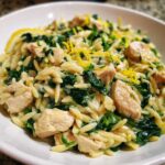 A close-up of a bowl filled with creamy chicken orzo with lemon and spinach, garnished with zest.