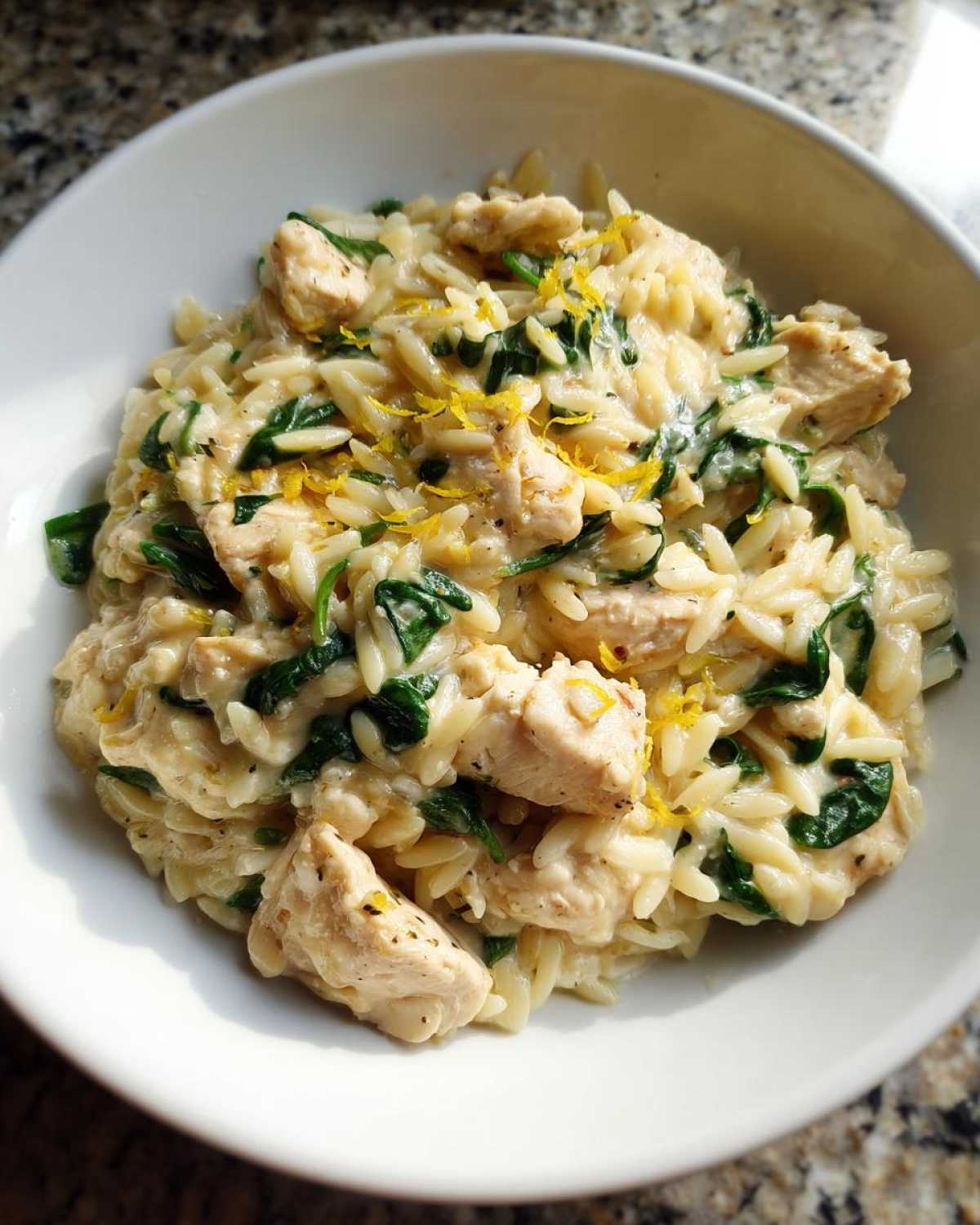 A close-up of a white bowl filled with creamy chicken orzo with lemon and spinach, topped with lemon zest.