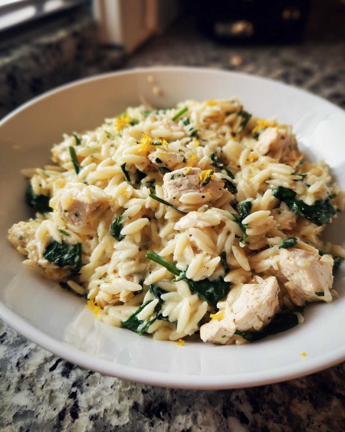 A close-up of creamy chicken orzo with lemon and spinach, topped with bright yellow lemon zest.