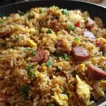 A close-up view of savory Creole Garlic Fried Rice featuring sausage slices, scrambled egg, and green onions in a dark skillet.