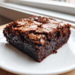 A single, rich, fudgy square of homemade brownies with a signature crackly top, served on a white plate.