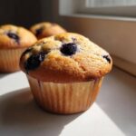 Close-up of a perfectly baked golden Blueberry Muffin with visible blueberries on top, set against natural window light.