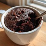 Close-up of a rich, dark Microwave Brownie in a Mug, topped with chocolate chips and being eaten with a spoon.