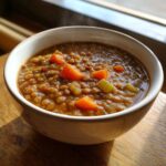 A close-up of a steaming white bowl filled with hearty One Pot Lentil Soup Cozy Dinner, showing lentils, carrots, and celery.