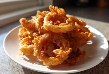 A close-up, featured image showing a mound of golden brown, crispy Onion Rings Extra Crispy piled high on a white plate.