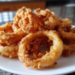 A close-up of a mound of golden-brown Onion Rings Extra Crispy piled high on a white plate.
