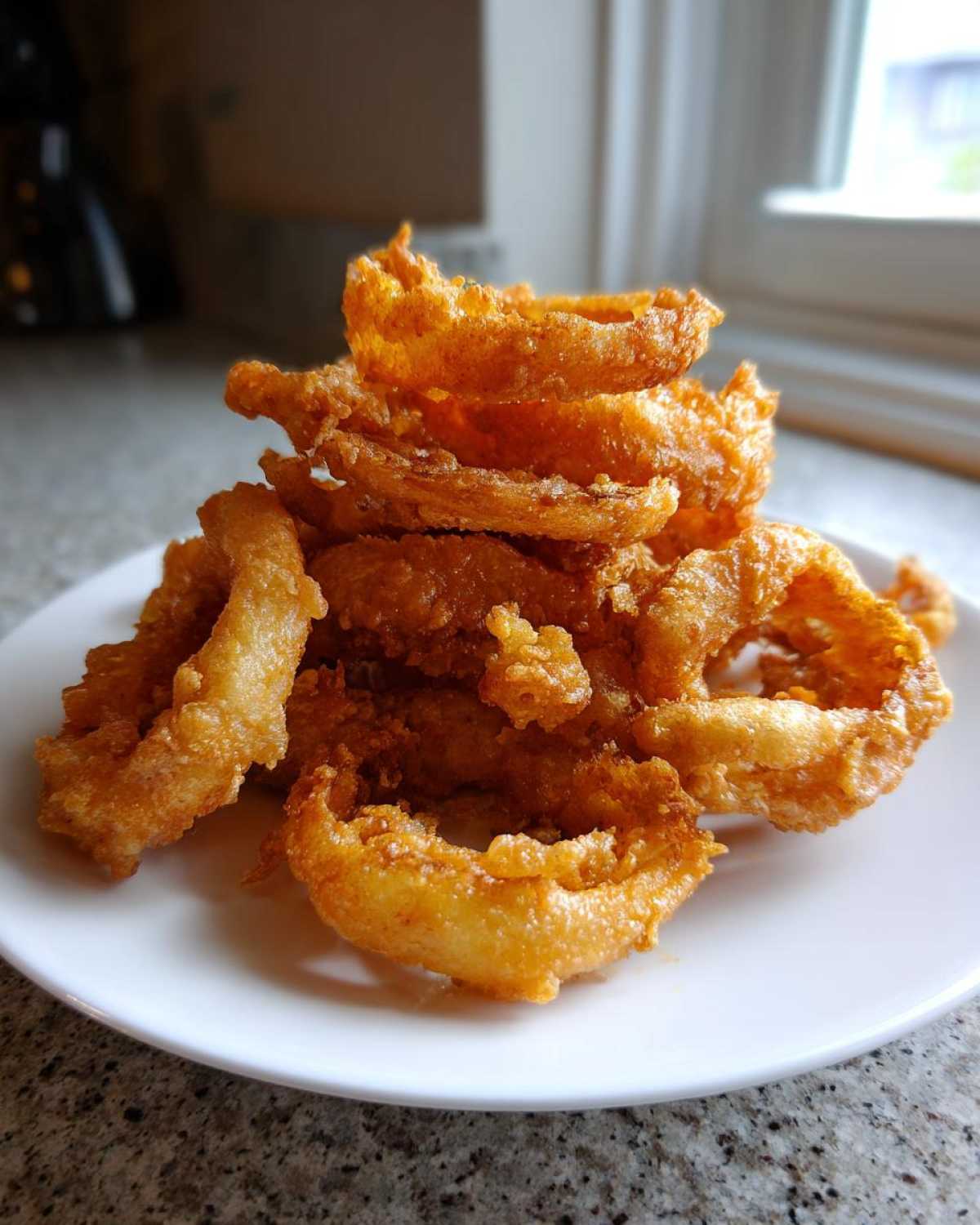 A close-up of a tall pile of golden-brown Onion Rings Extra Crispy stacked on a white plate.