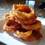 A close-up stack of golden brown Onion Rings Extra Crispy piled high on a white plate.
